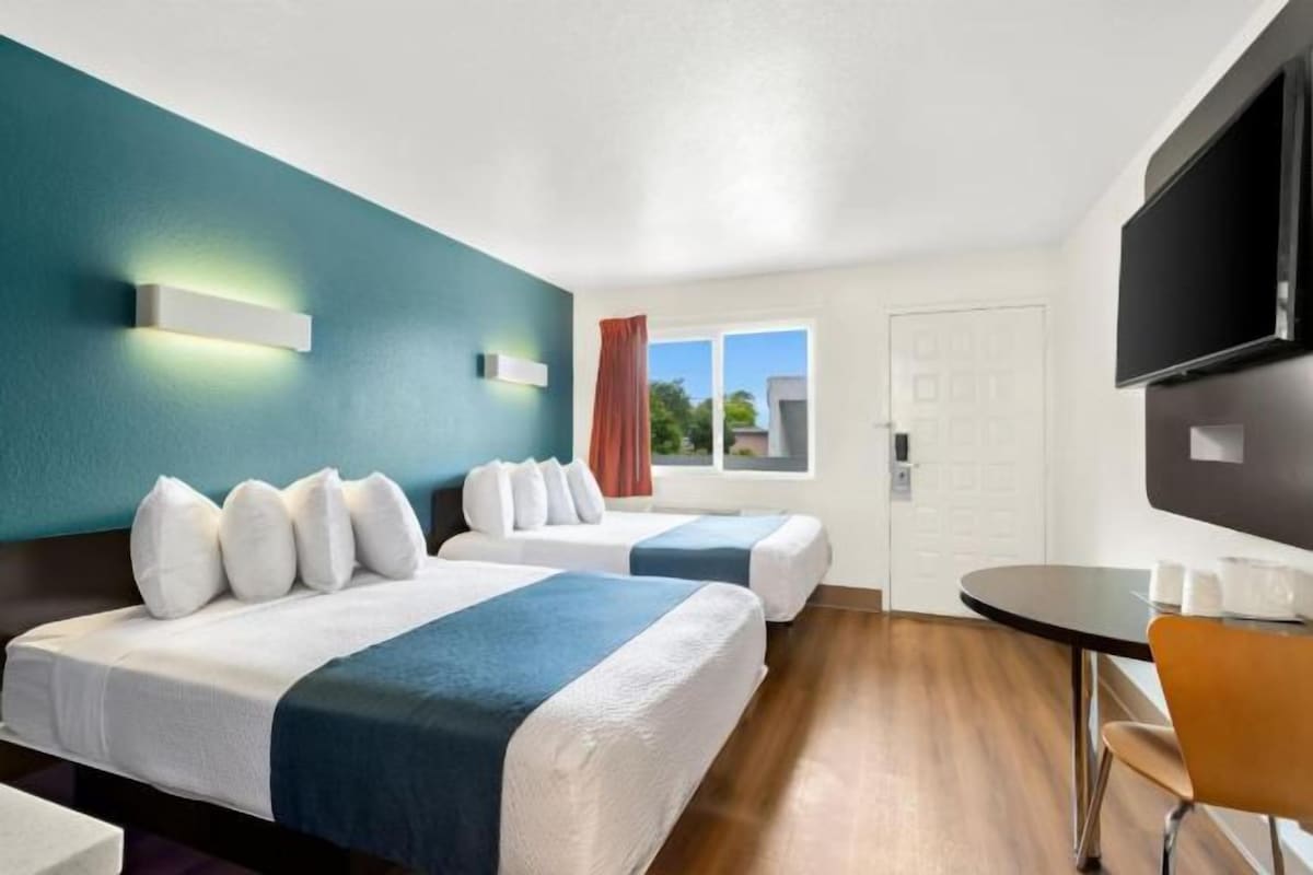 Quarto de hotel com duas camas, televisão, janela com cortinas e uma mesa pequena com chávenas.