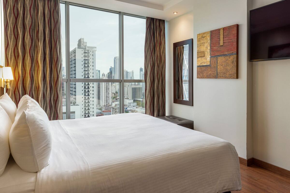 A hotel room with a large bed, a city view, a flat-screen TV, and a modern art piece on the wall.
