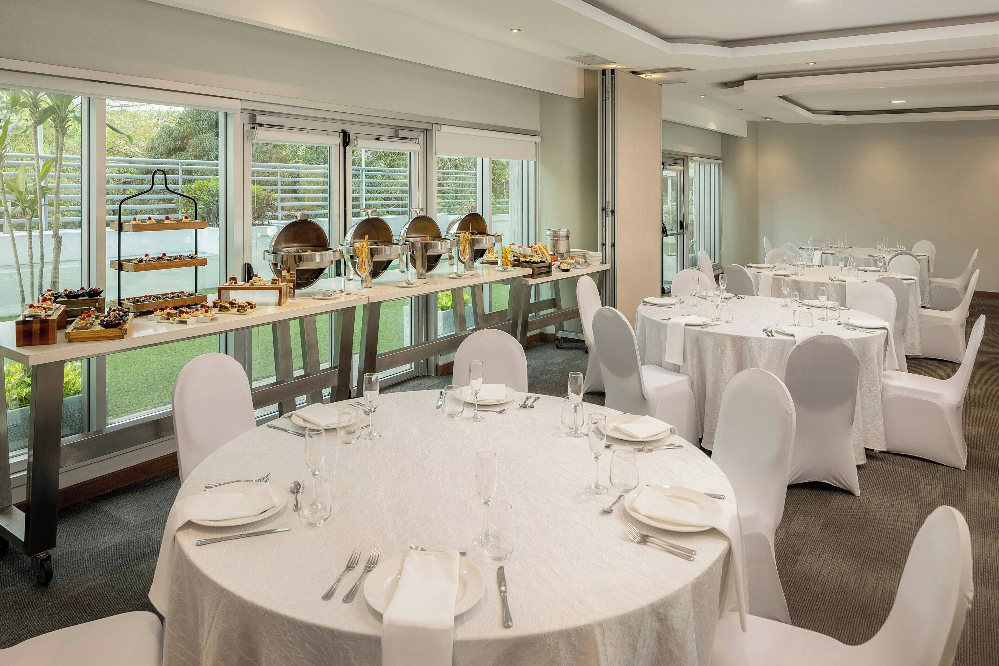 A banquet hall with round tables, white chairs, and a buffet area with food and drink.