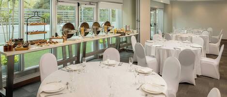 A banquet hall with round tables, white chairs, and a buffet area with food and drink.