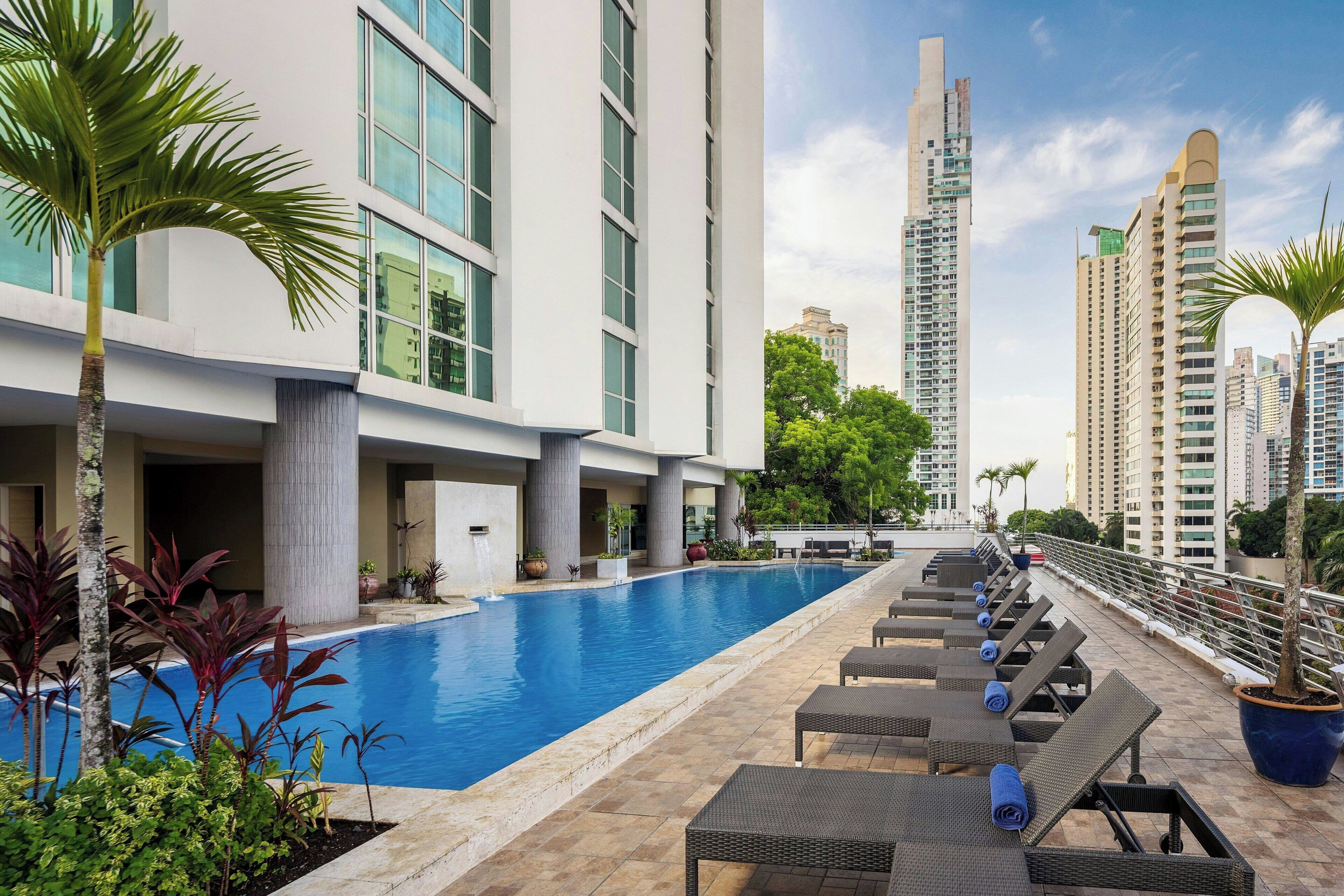 A swimming pool with lounge chairs, a palm tree, and tall buildings in the background.