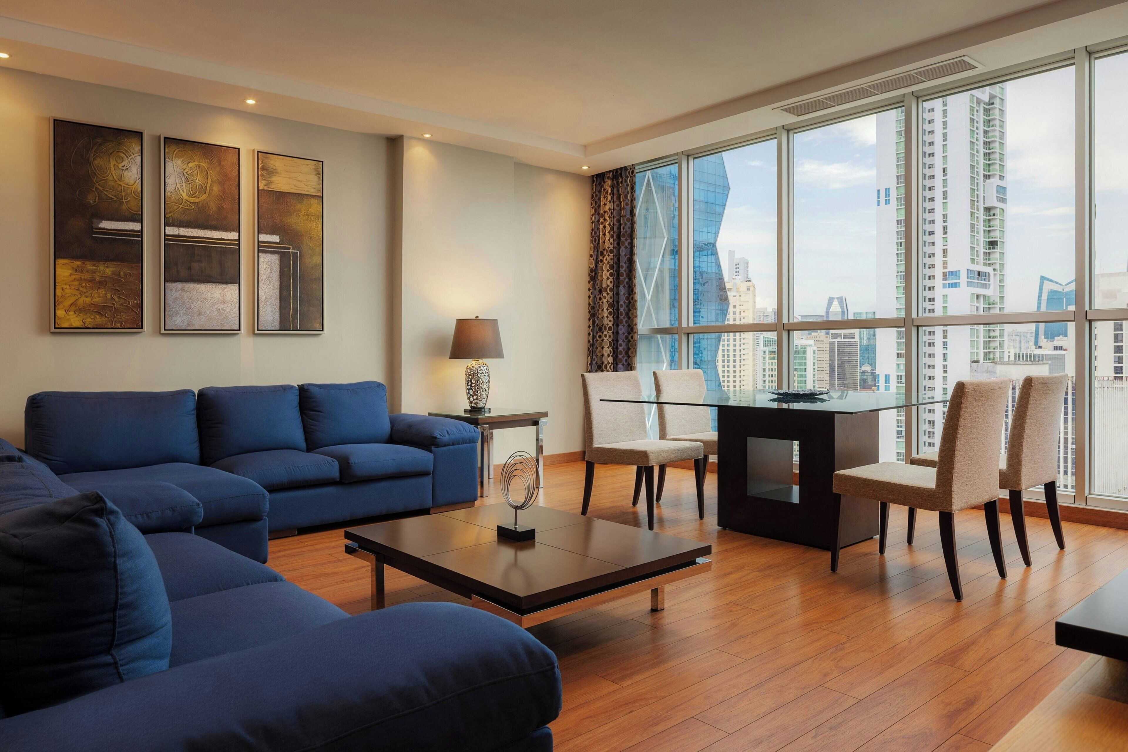 A modern living room with a blue sofa set, a glass coffee table, and a dining area with chairs and a table. Large windows offer a view of the cityscape.
