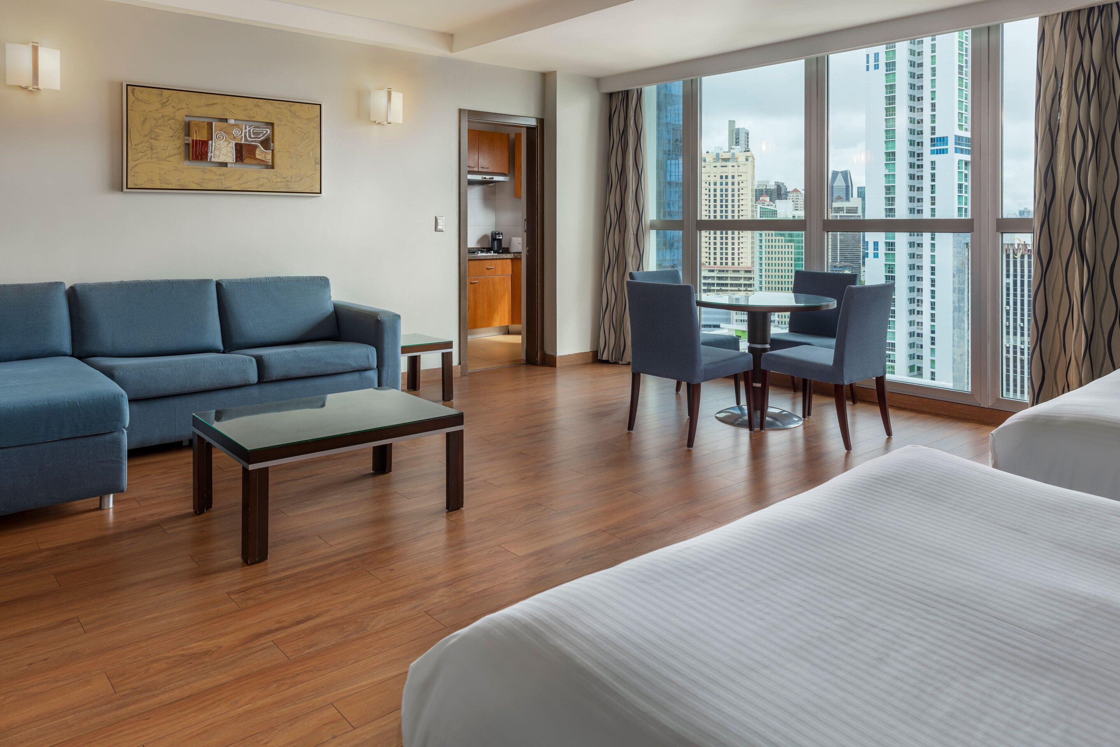Habitación de hotel con sofá, una mesita, un comedor con sillas, cama, ventana con vista a la ciudad y una zona de cocina.