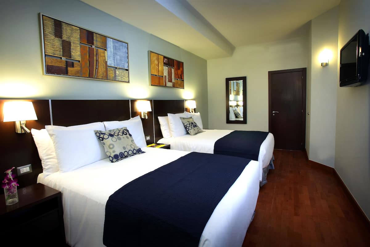 A hotel room with two beds, nightstands, wall-mounted lamps, a flat-screen TV, and artwork on the walls.