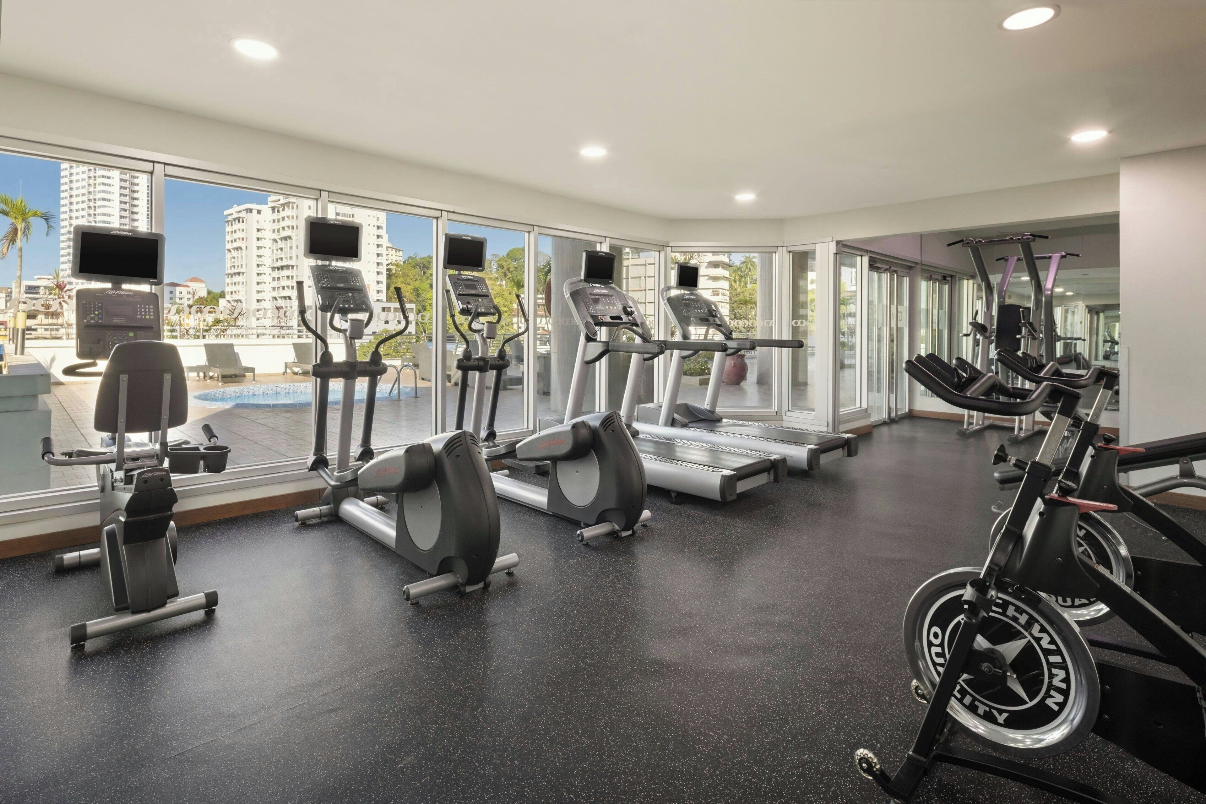 Un gimnasio con cintas de correr, elípticas y una máquina de remo, ventanas amplias y vista a edificios.