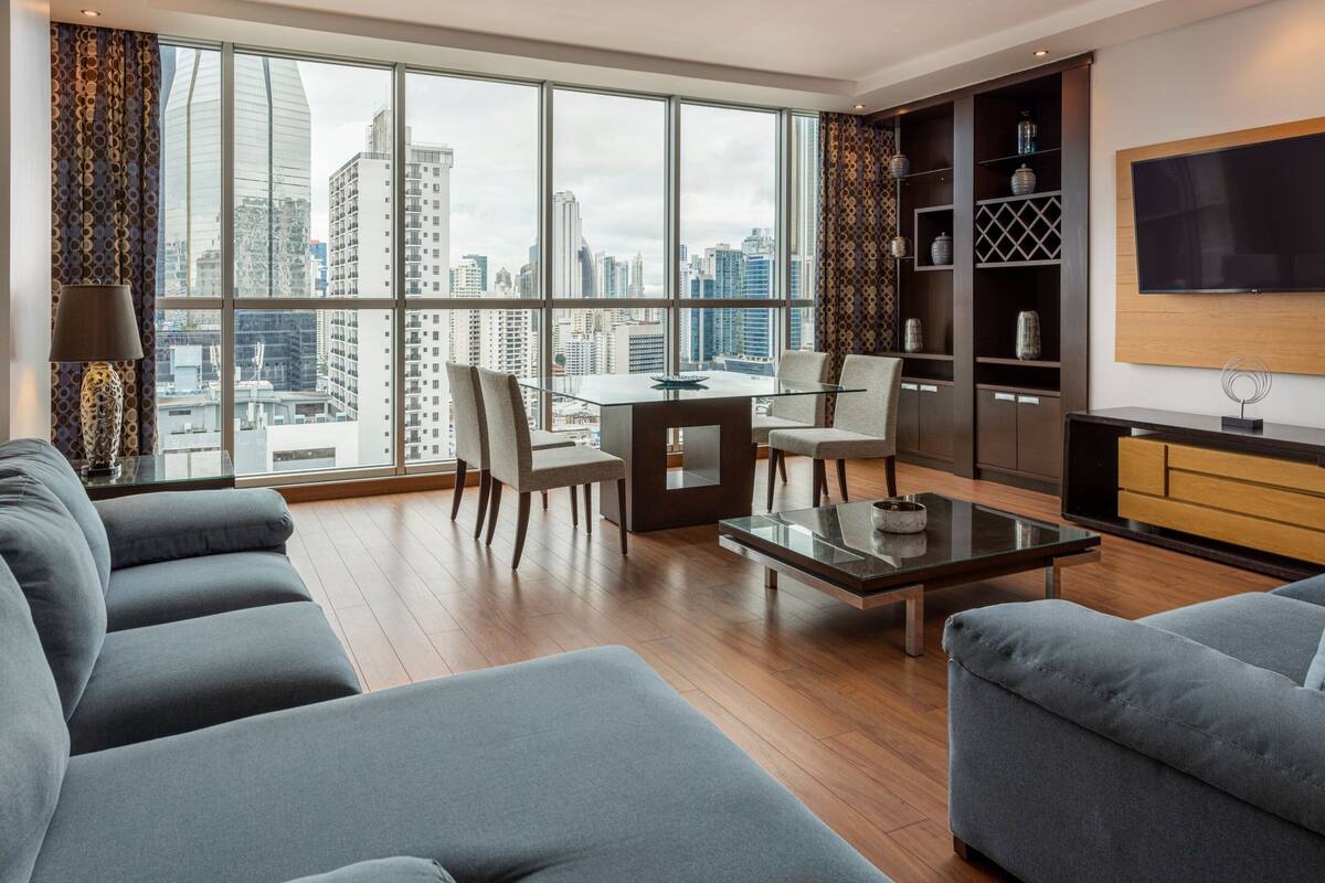 A modern living room with a city view, a dining table, and a TV mounted on the wall.