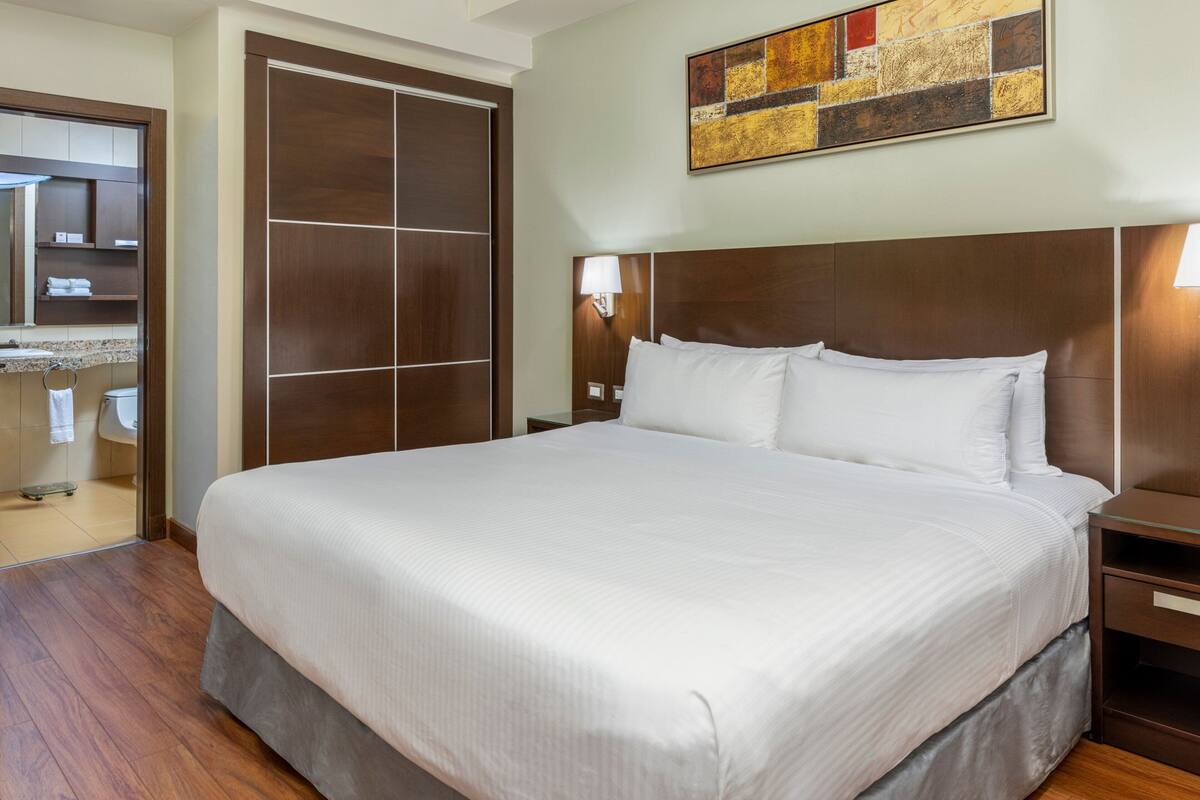 A hotel room with a large bed, wooden headboard, a bedside table, a nightstand, a mirror, a bathroom with a toilet and sink, and a wall-mounted artwork.