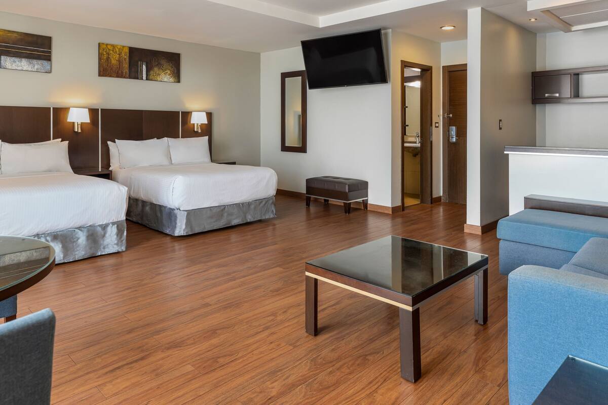 A hotel room with two beds, a small table, a sofa, a TV, and a kitchenette.