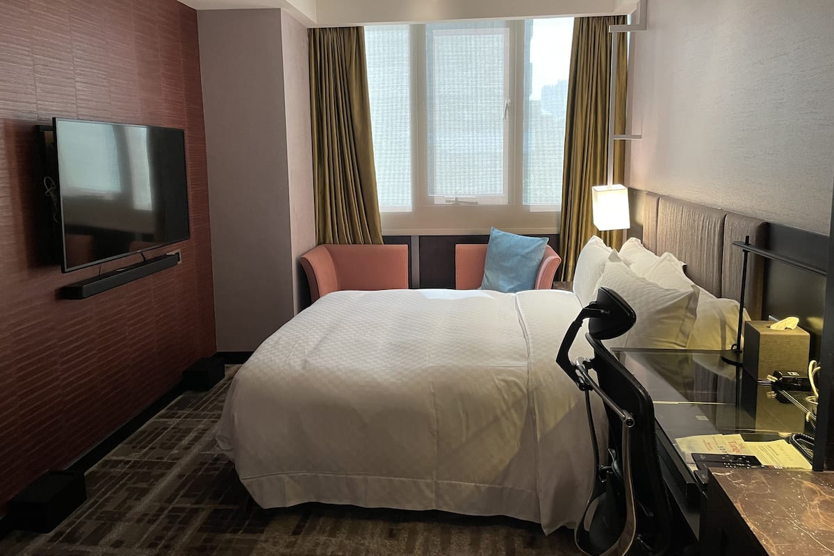 Une chambre d’hôtel dotée d’un grand lit, d’un téléviseur à écran plat fixé au mur, d’un bureau avec une lampe et d’une fenêtre avec des rideaux.