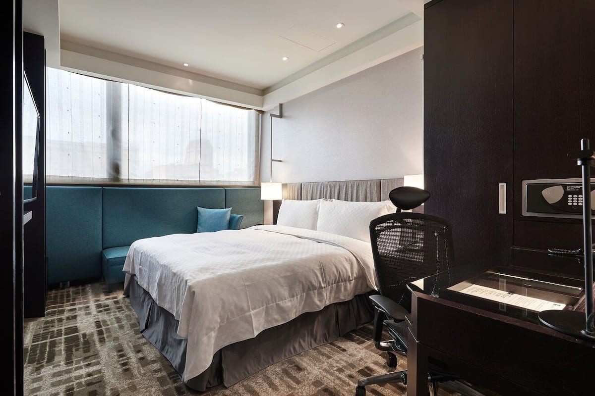 Une chambre d’hôtel comprenant un lit, un bureau, une chaise, une fenêtre avec des stores et une armoire de couleur sombre.