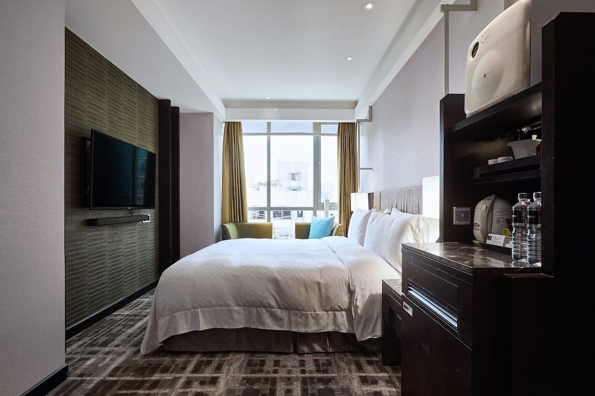 Une chambre d’hôtel dotée d’un grand lit, d’un téléviseur à écran plat fixé au mur, d’un minibar et offrant une vue sur la ville grâce à de grandes fenêtres.
