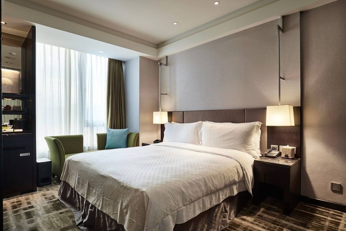 Une chambre d’hôtel avec un grand lit, deux lampes de chevet, une table de nuit, un fauteuil vert et une vue sur l’extérieur à travers des rideaux légers.