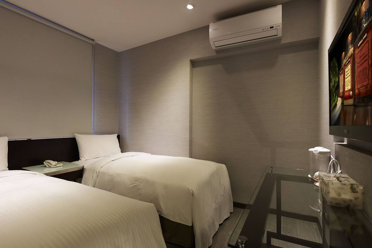 A hotel room with two beds, a nightstand with a phone, an air conditioning unit, a small table with a towel, and a mirror with a red sign.