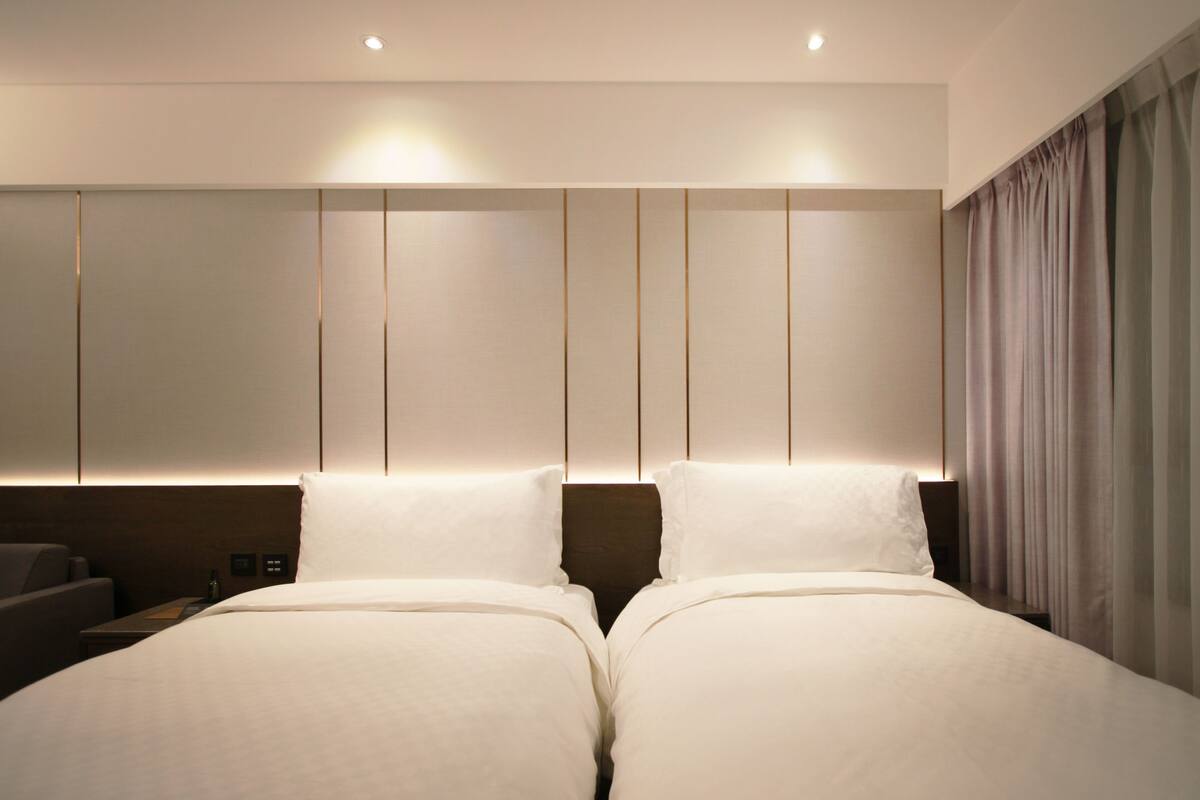 Ein Hotelzimmer mit zwei Betten, einem Kopfteil mit vertikalen Linien, einem Nachttisch und einem Vorhang auf der rechten Seite.