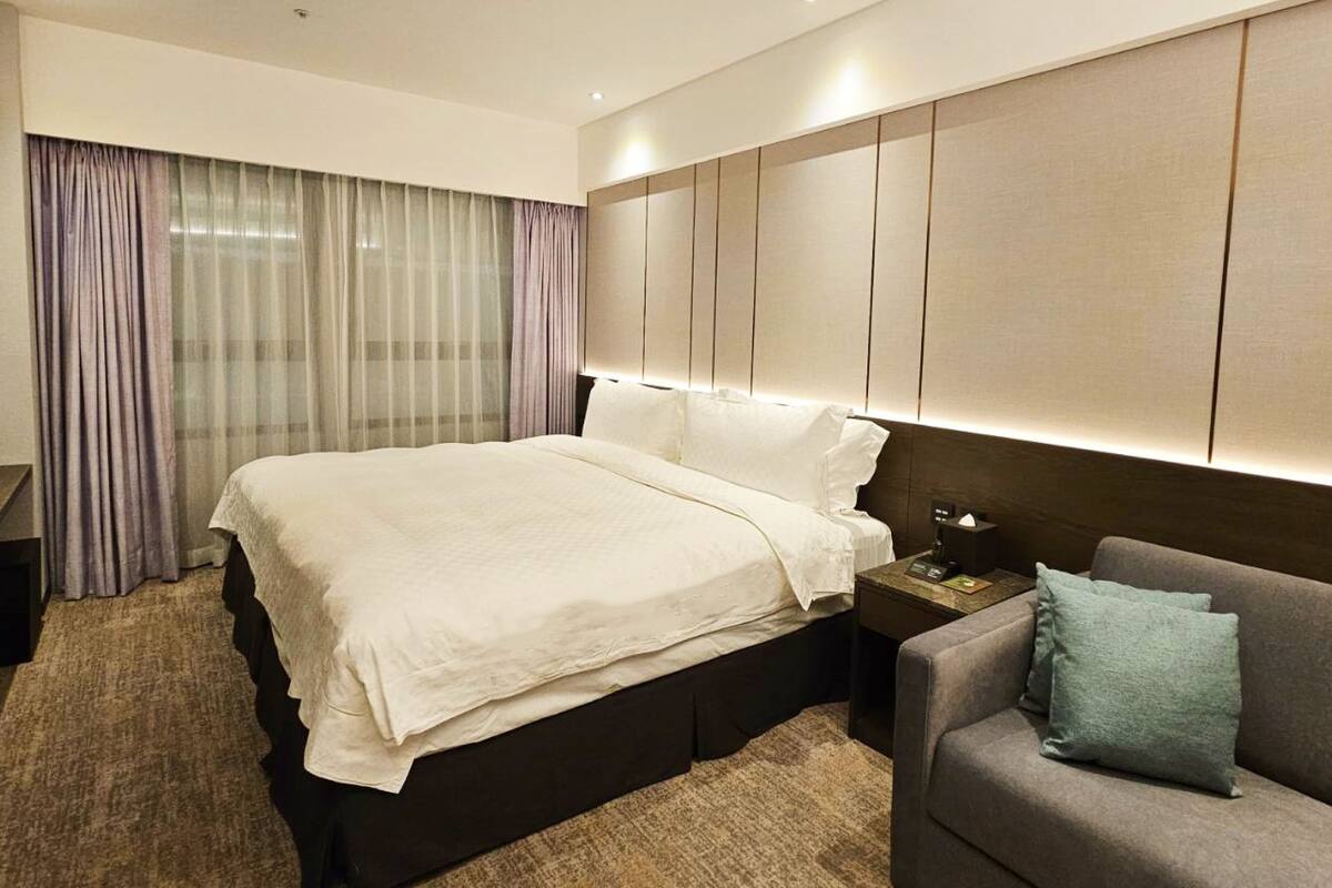 Ein modernes Hotelzimmer mit einem großen Bett, einem Nachttisch mit Lampe, einem grauen Sofa mit Kissen und einem großen Fenster mit Vorhängen.