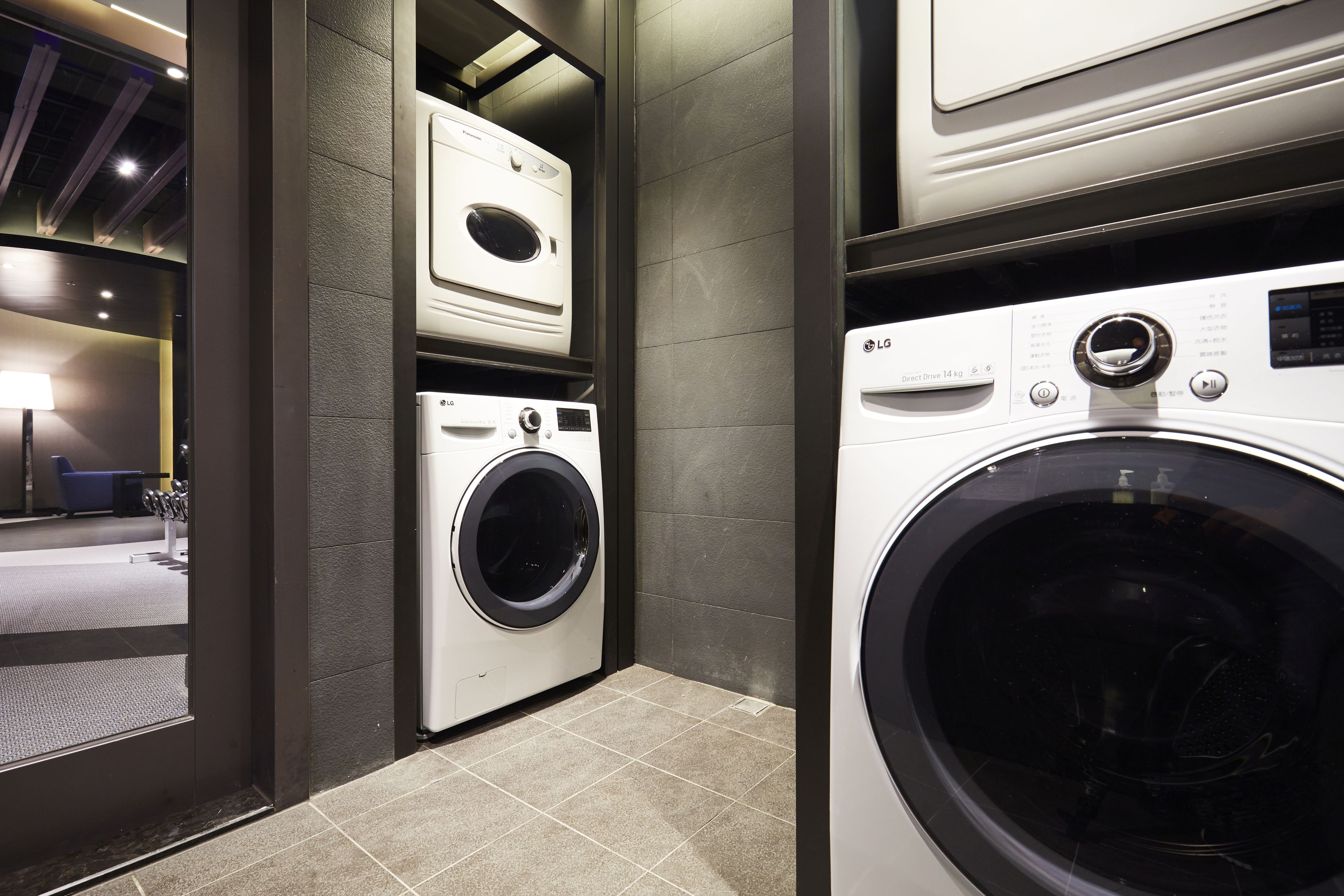 A laundry room with two washing machines, a dryer, and a small shelf with a lamp.