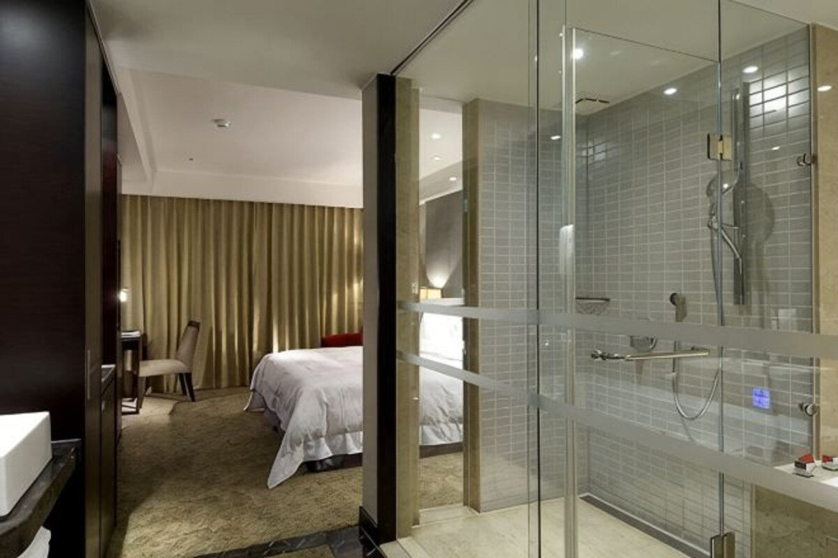 A hotel room with a bed, a chair, a desk, and a glass-enclosed shower area.