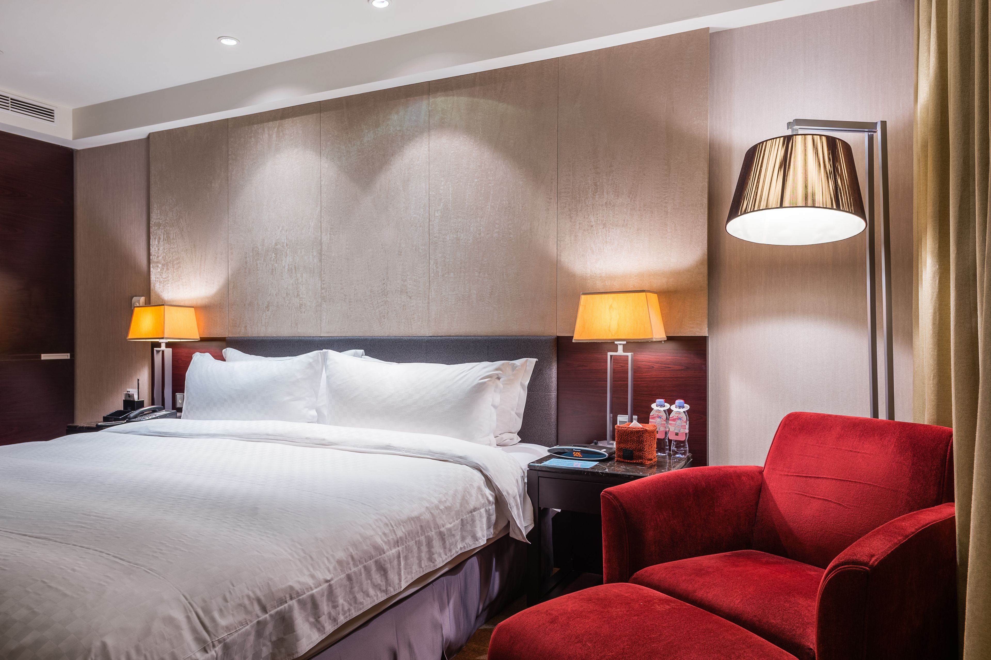 A hotel room with a large bed, two bedside lamps, a red armchair, and a floor lamp.
