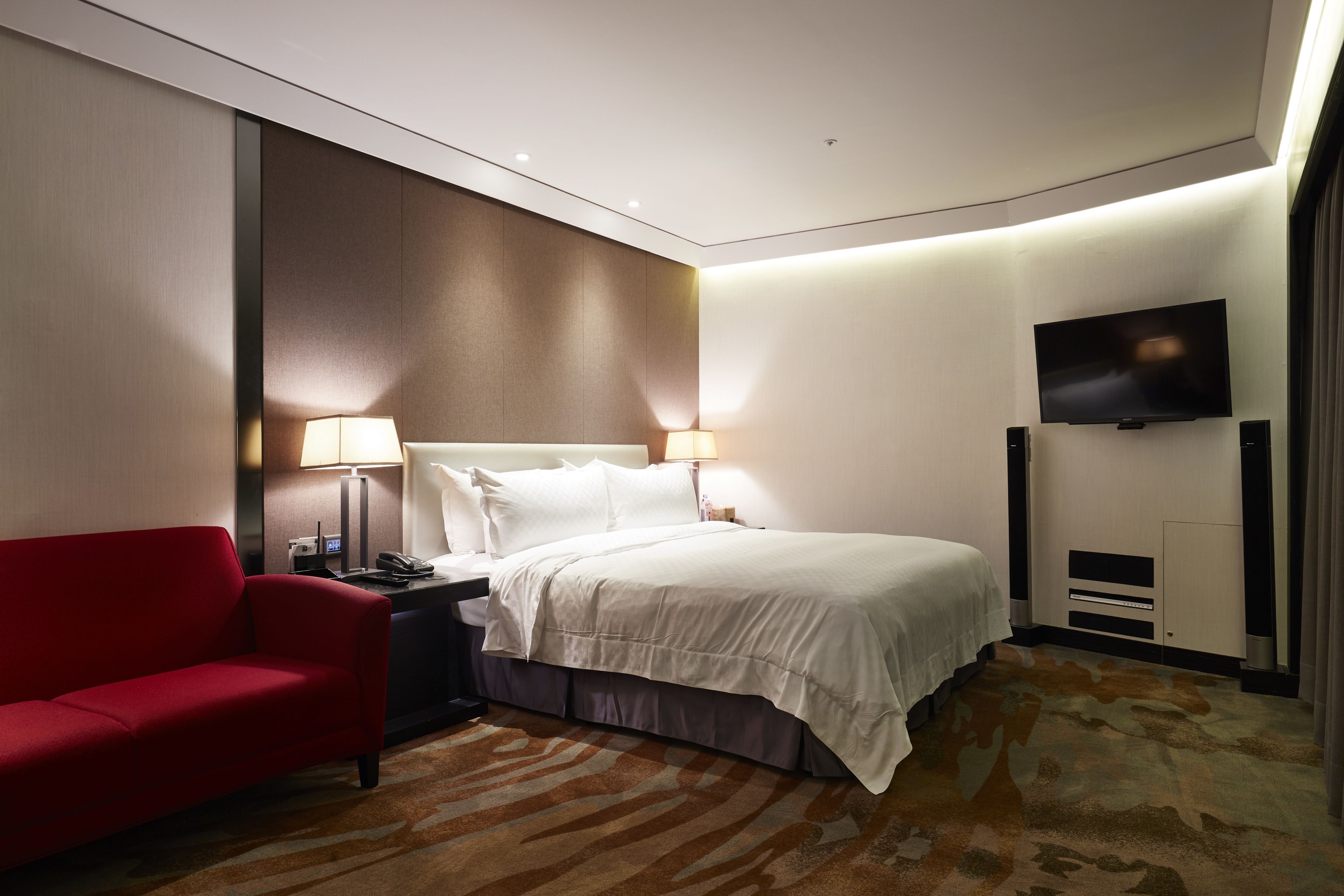 A hotel room with a bed, a red sofa, a TV, and a nightstand with a lamp.