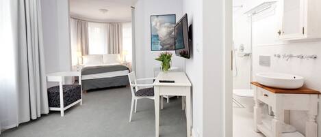 A hotel room with a bed, a desk, a chair, a TV, and a bathroom with a sink and mirror.
