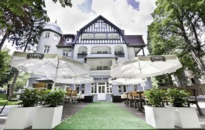A hotel with a traditional European architectural style, featuring a central courtyard with a green carpet and white umbrellas labeled "ZYWIEC VILLA POLANICA".