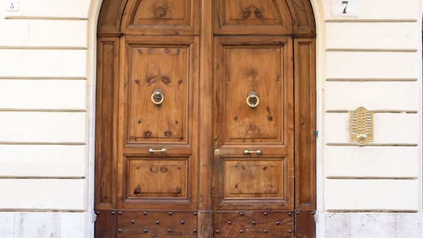 A double wooden door with a metal handle, flanked by white columns and a balcony with balustrades. - Domus RomAntica (Rome)
