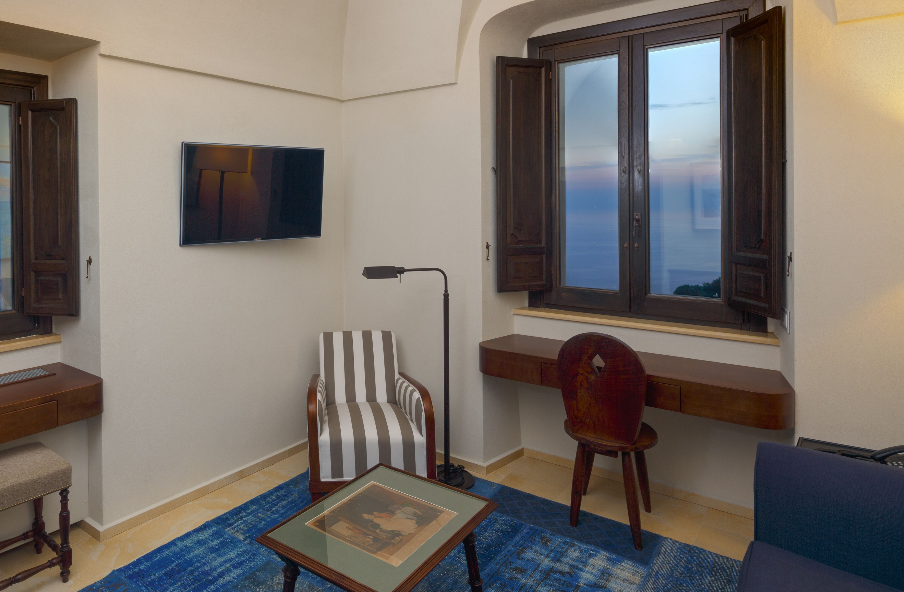 A room with a large window, a flat-screen TV mounted on the wall, a striped armchair, a red chair, a small table with a picture, and a blue rug.