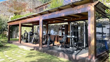 Un gimnasio al aire libre con equipamiento de ejercicio, una zona de estar techada y un muro de piedra al fondo.
