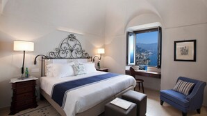 A hotel room with a bed, bedside tables, a chair, a desk, and a window with a view. - Monastero Santa Rosa Hotel & Spa (Conca dei Marini)