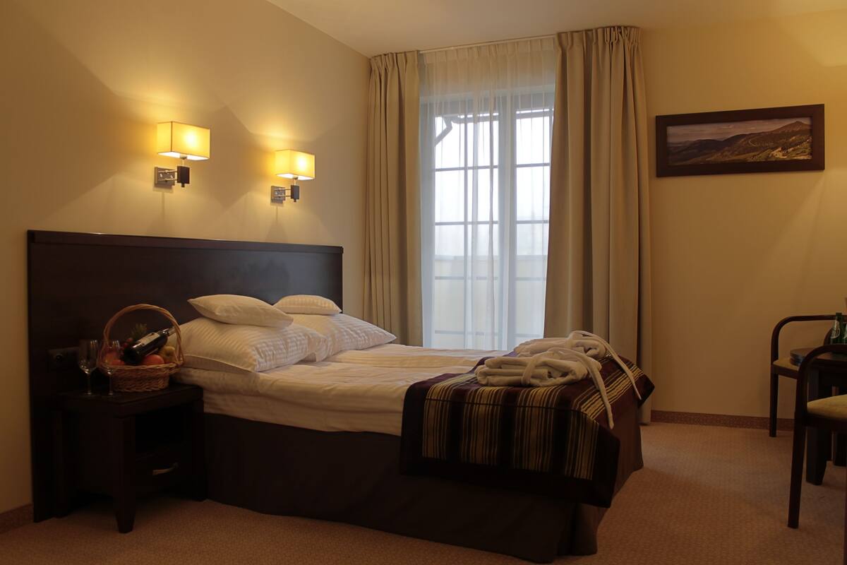 A hotel room with a large bed, two bedside lamps, a window with curtains, a framed picture on the wall, and a small table with a basket and a bottle.