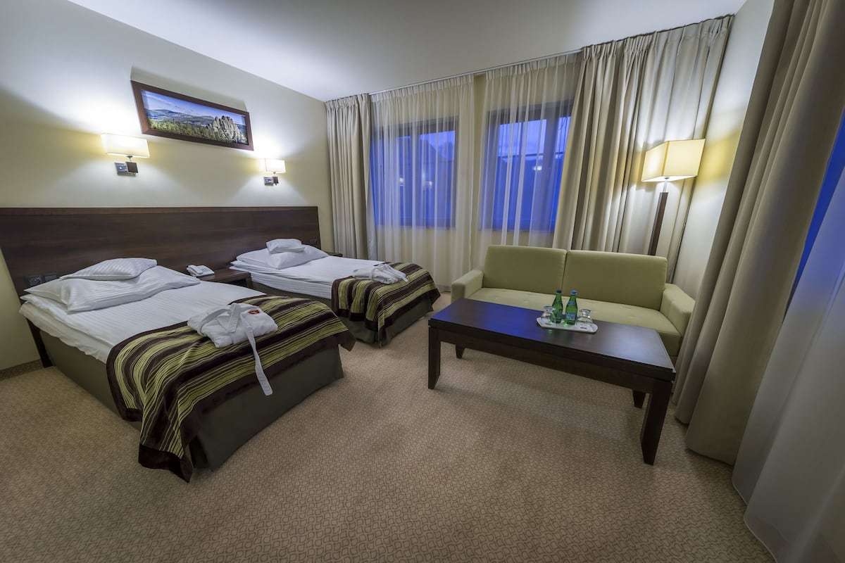 A hotel room with two beds, a sofa, a small table, and a view of the outdoors through sheer curtains.