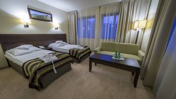 Habitación de hotel con dos camas, un sofá, una mesita y vistas al exterior a través de cortinas traslúcidas.