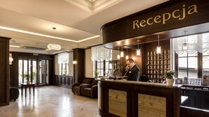 A reception area with a wooden reception desk, a receptionist, and a seating area with leather chairs. - Hotel Krysztal Conference & Spa (Szklarska Poreba)