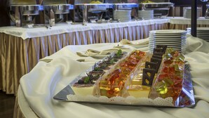 A buffet setup with various dishes and desserts on trays, a table with stacked plates, and a backdrop of a room with curtains and a patterned wall. - Hotel Krysztal Conference & Spa (Szklarska Poreba)