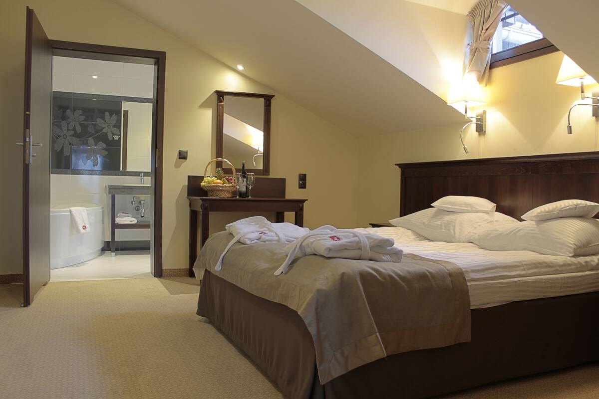 A bedroom with a bed, a bedside table with a lamp, a window, and a bathroom visible through an open door.