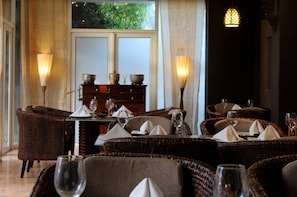 Comedor con sillas de mimbre, mesas dispuestas con servilletas blancas y copas de vino, y una ventana con vistas a la vegetación.