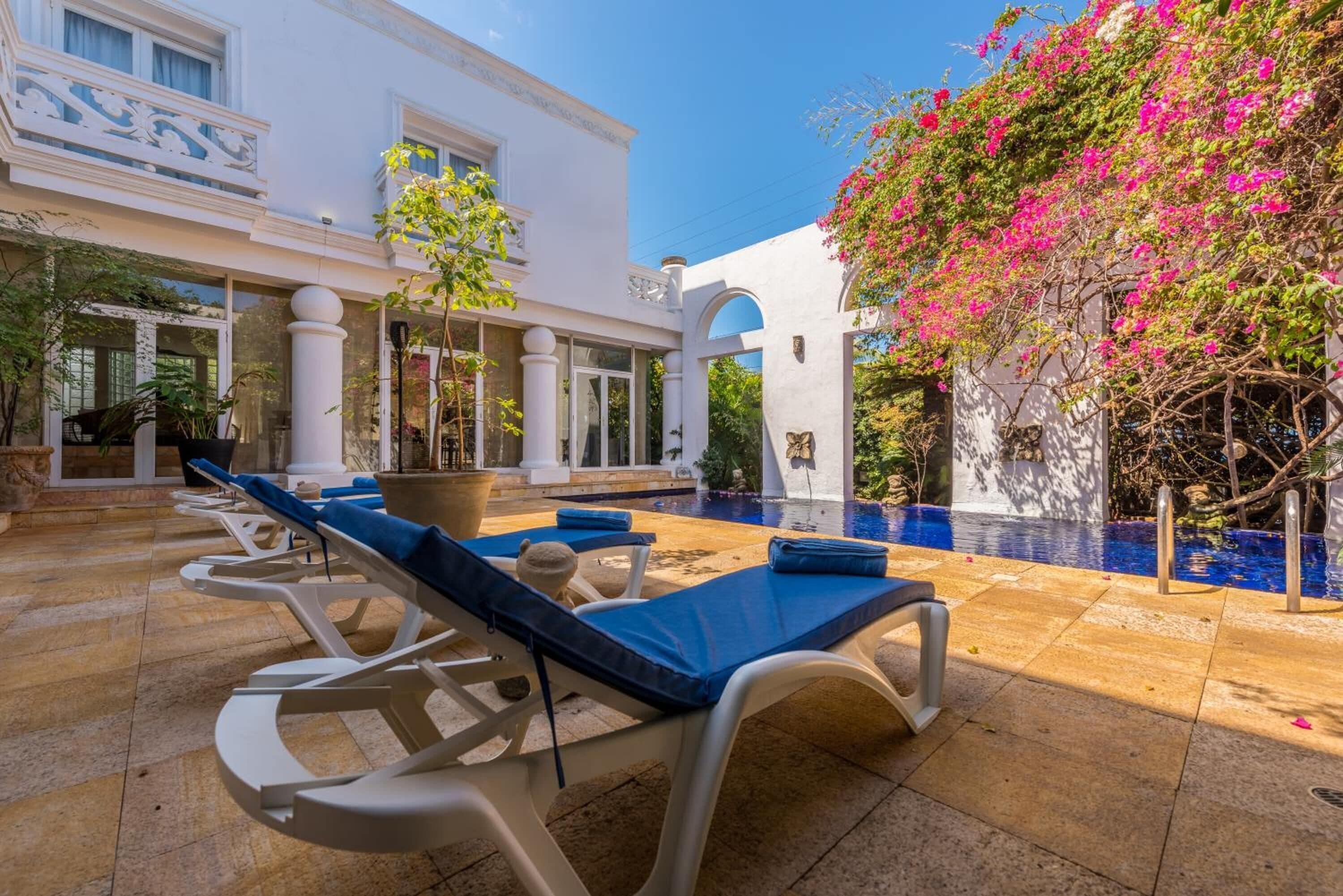 Zona de piscina con sillones de descanso, una piscina y un edificio con columnas y arcos.