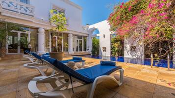 Zona de piscina con sillones de descanso, una piscina y un edificio con columnas y arcos.