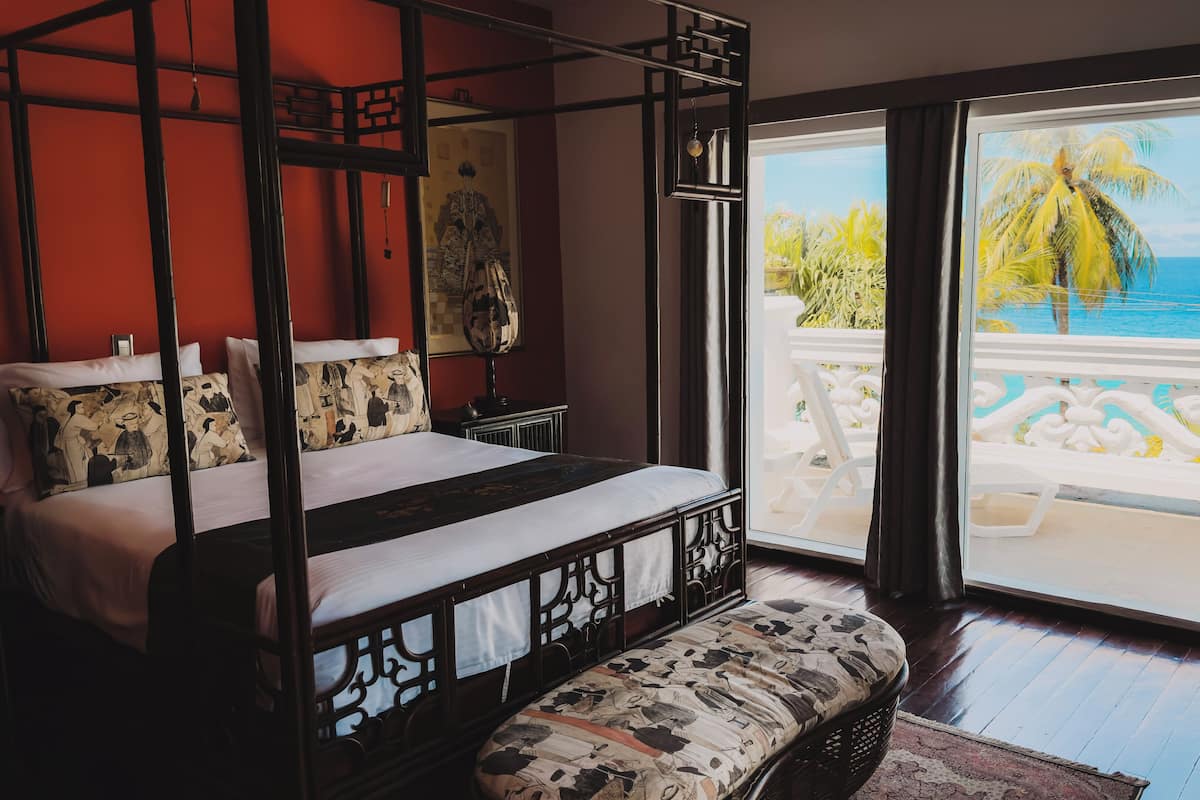 Een slaapkamer met een hemelbed, een bankje en uitzicht op een zwembad en palmbomen.