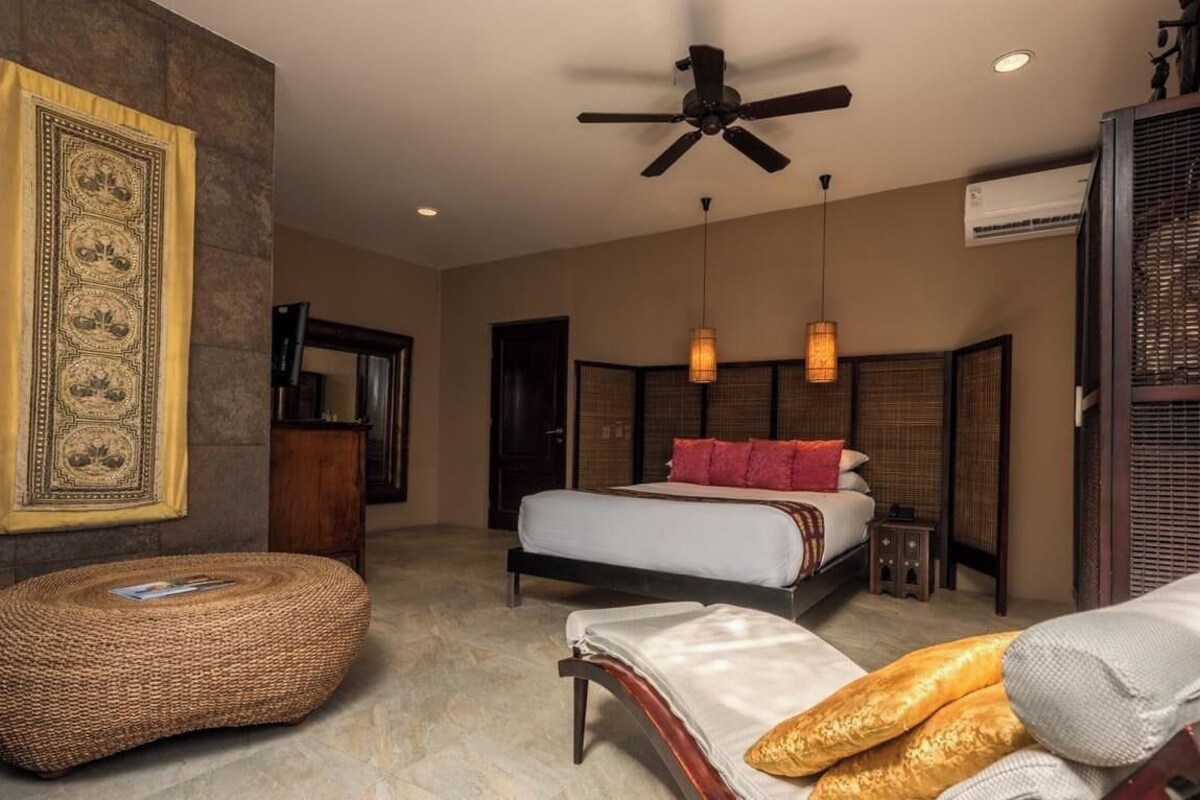 Une chambre à coucher comprenant un lit, un ventilateur de plafond, un pouf en osier et un climatiseur fixé au mur.
