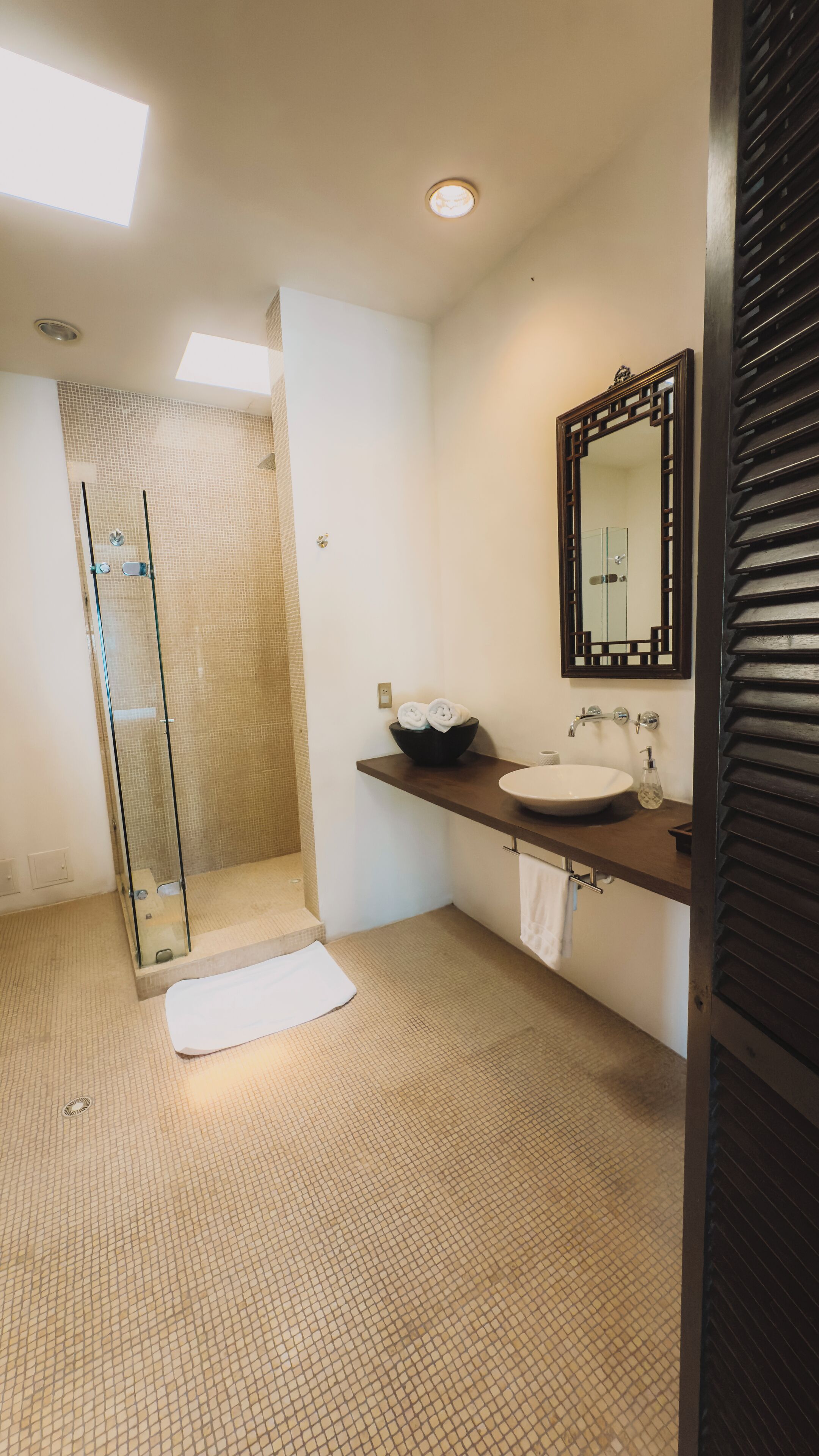 Un baño moderno con cabina de ducha de vidrio, un espejo grande y suelo de baldosas color beige.