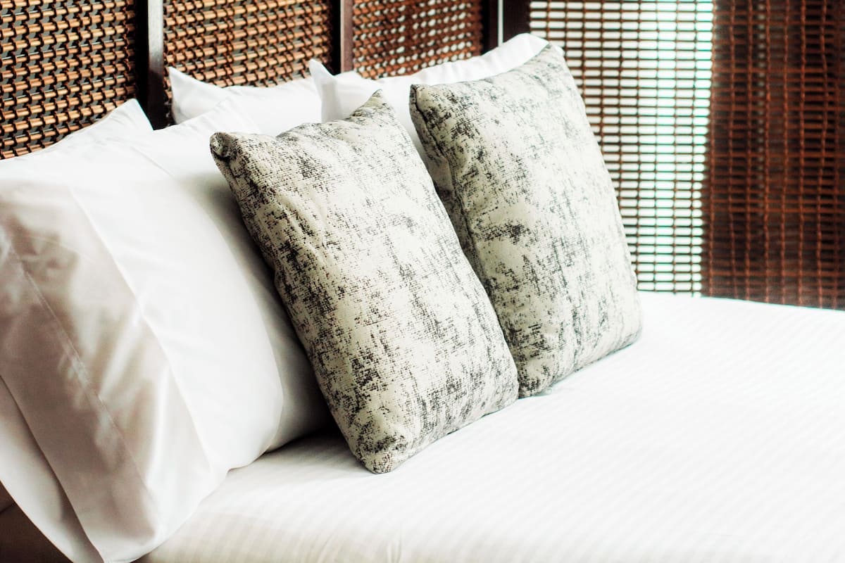 Een bed met witte beddengoed en twee gestreepte kussens, een donker houten nachtkastje met decoratie, en een geweven hoofdbord.