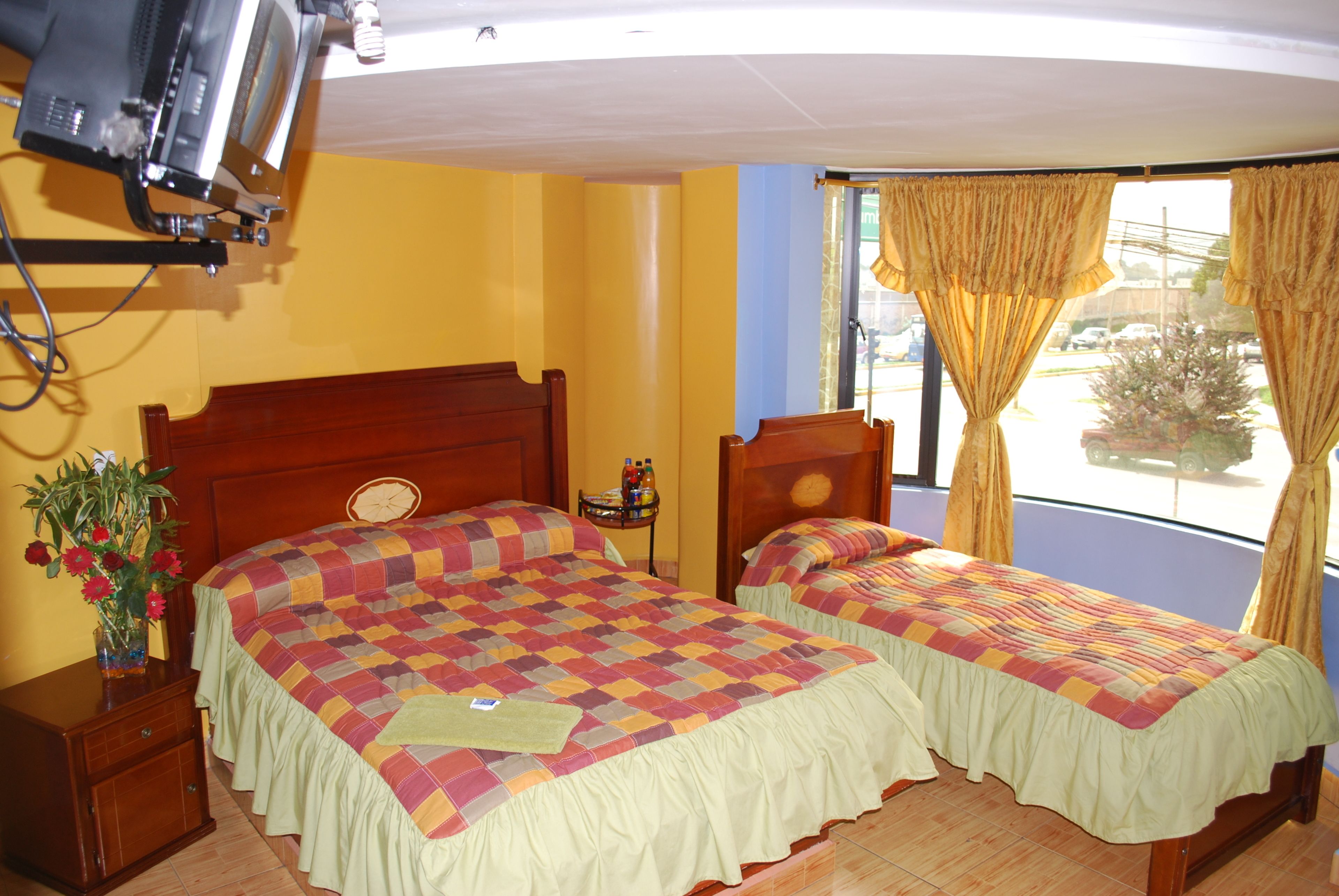 A hotel room with two beds, a television mounted on the wall, a small table with a vase of flowers, and a window with curtains.