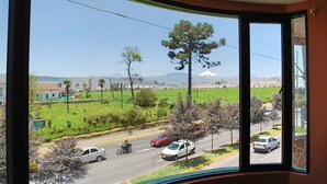A view from a window overlooking a road with cars and a cyclist, a green field, and a mountain in the distance. - Hotel Gran Quitumbe (Quito)