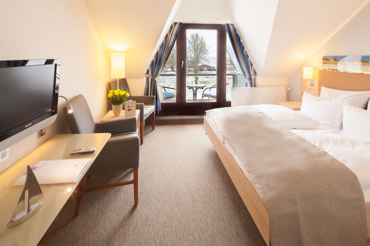 A hotel room with a bed, a desk with a TV, a chair, a table with a lamp, and a view of the outdoors.