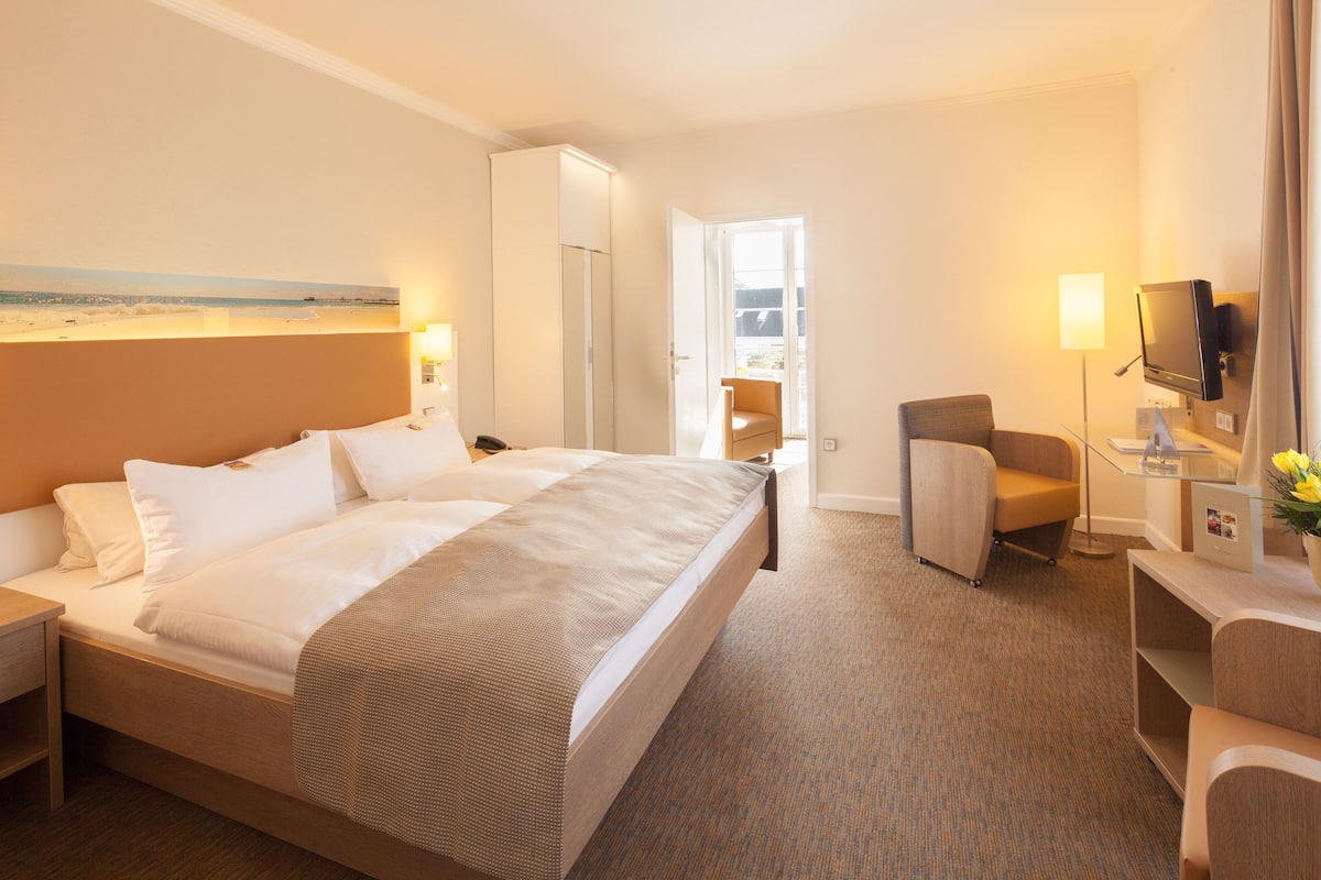 A hotel room with a large bed, a desk, a chair, a TV, and a window with a view of the beach.