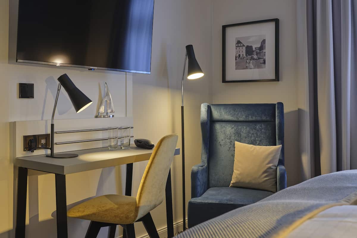 A hotel room with a bed, a desk with a computer, a chair, a TV mounted on the wall, a floor lamp, and a framed picture.