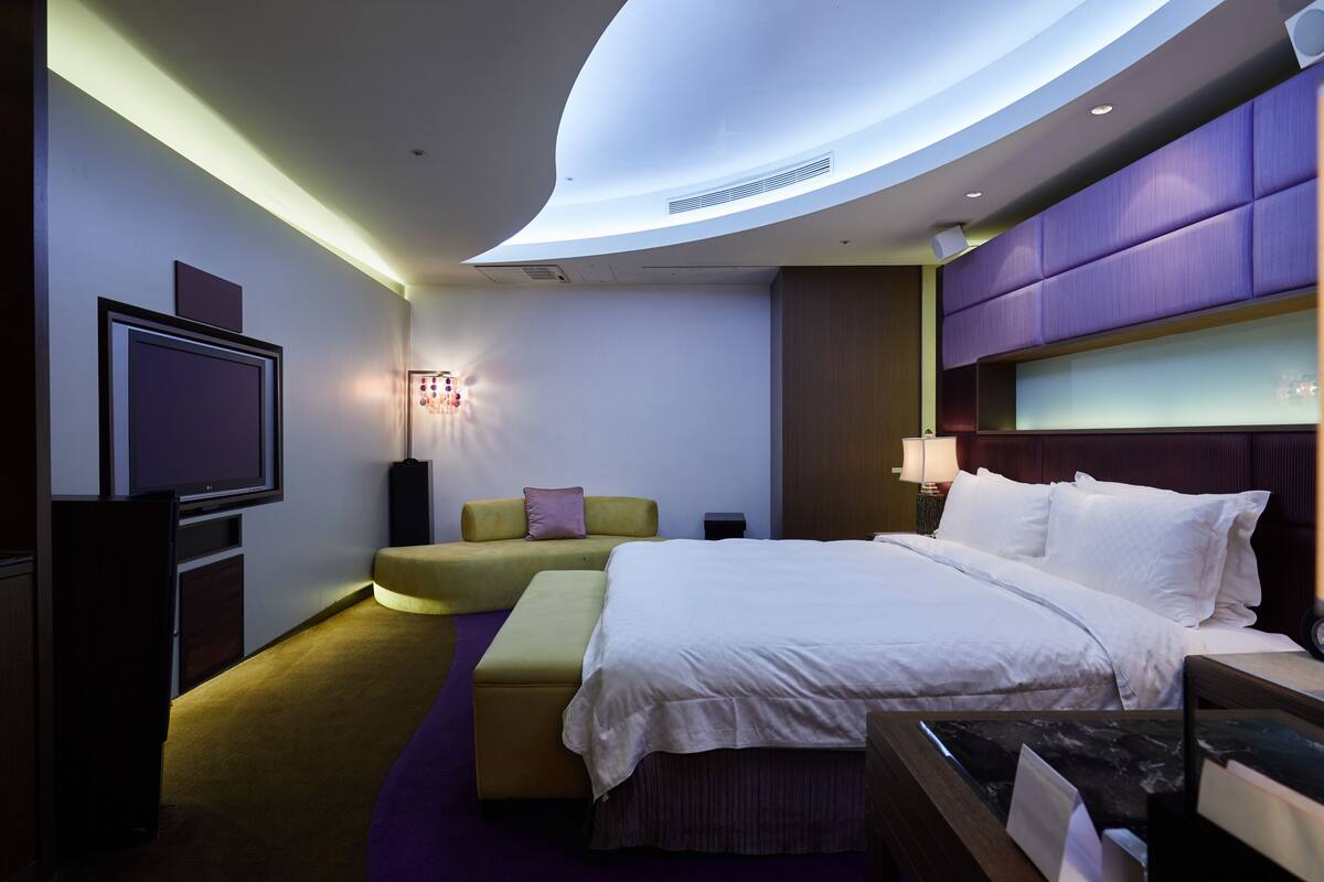 A modern hotel room with a curved ceiling, a large bed, a seating area, and a television.