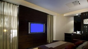 Una habitación de hotel con una cama, un televisor montado en un panel de madera oscura, un escritorio con una lámpara y un espejo grande.