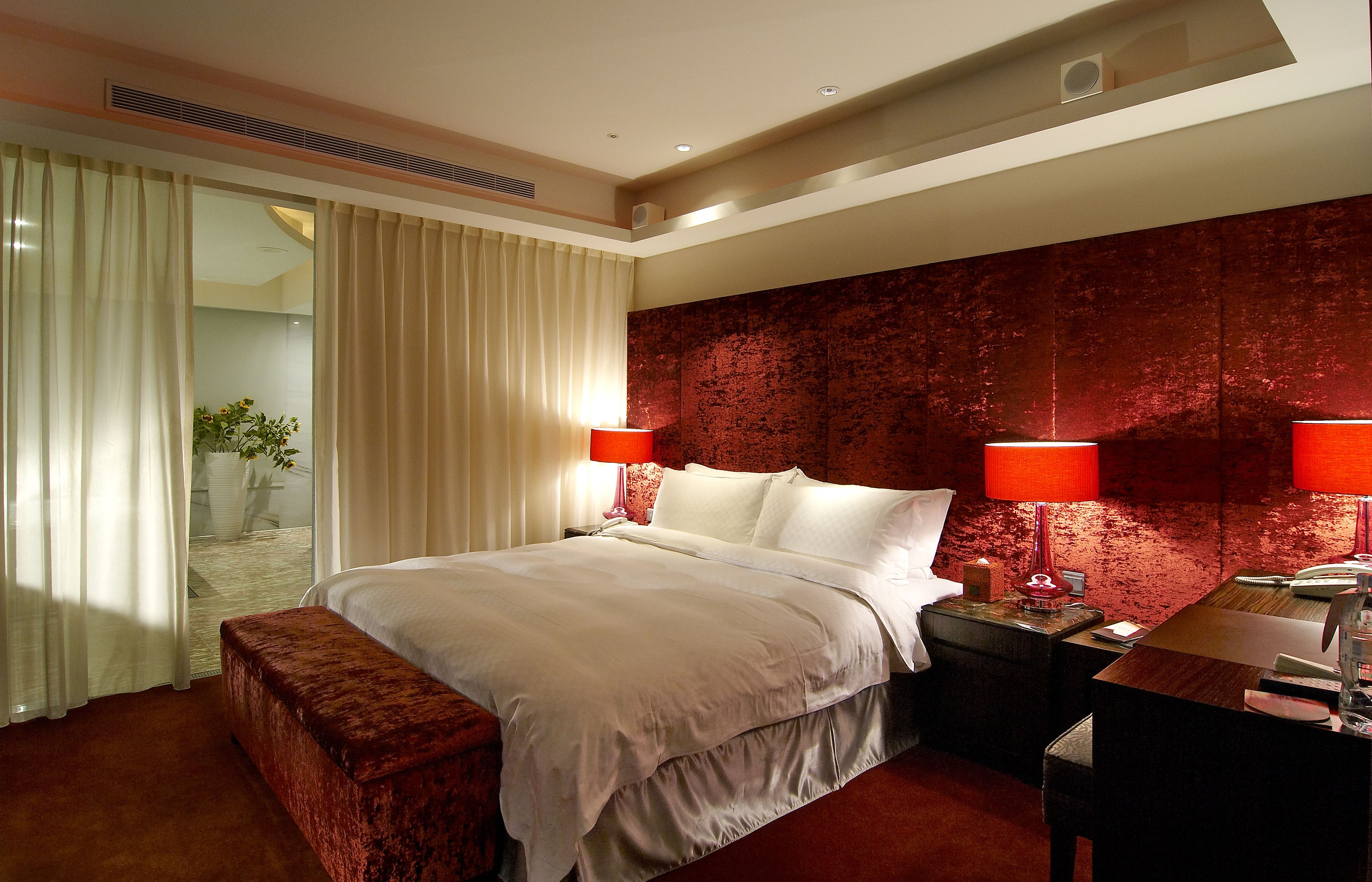 A hotel room with a large bed, two bedside lamps, a wooden nightstand, a desk, and a red accent wall.